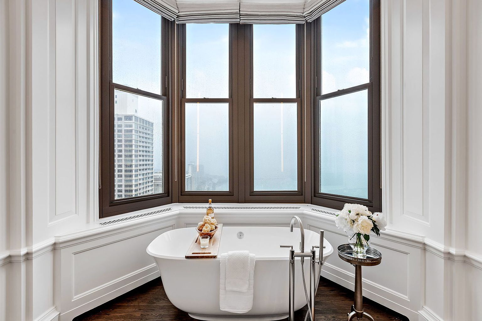 This is a luxurious primary bathroom featuring a freestanding soaking tub positioned in front of a large bay window with views of the city skyline. The room is decorated with white wainscoting and dark hardwood floors, creating a sophisticated and relaxing atmosphere. A small side table with a vase of white flowers adds a touch of elegance.