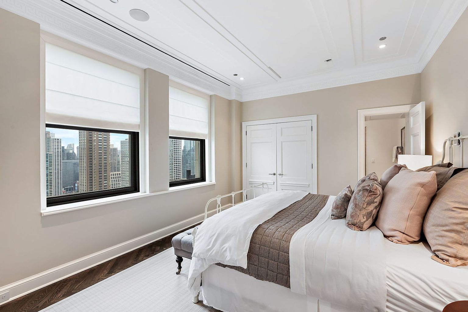 This is a well-lit primary bedroom featuring two large windows with city views. The room is decorated in neutral tones with a white bedspread and a brown quilted throw. The architectural details include detailed ceiling molding and dark hardwood floors, creating a sophisticated and comfortable atmosphere.