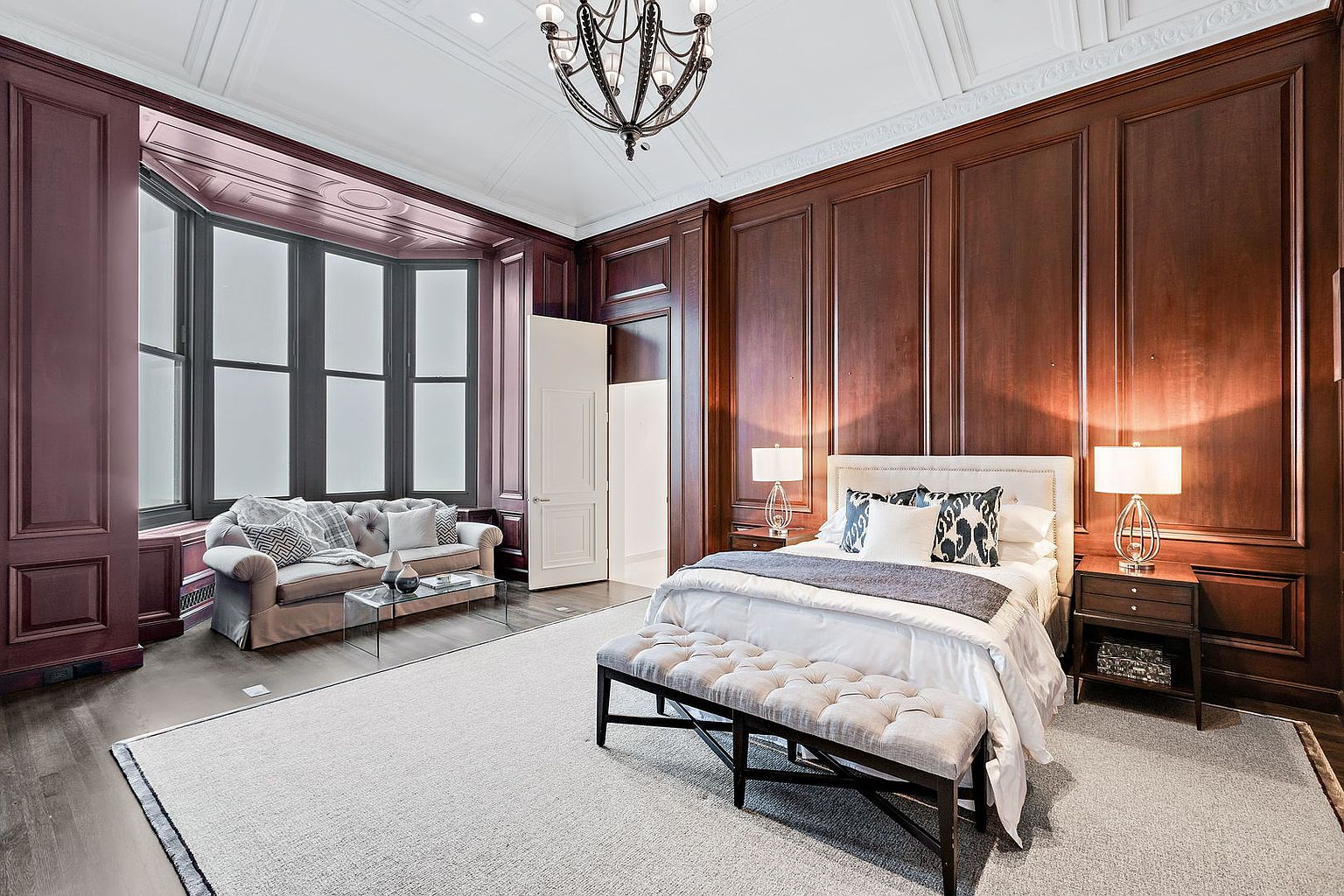 This is a luxurious primary bedroom featuring rich wood paneling, a large bay window with a seating area, and a plush bed with a tufted bench. The room is illuminated by table lamps on either side of the bed and a chandelier, creating a warm and inviting atmosphere. The overall style is elegant and sophisticated, perfect for a high-end real estate listing.