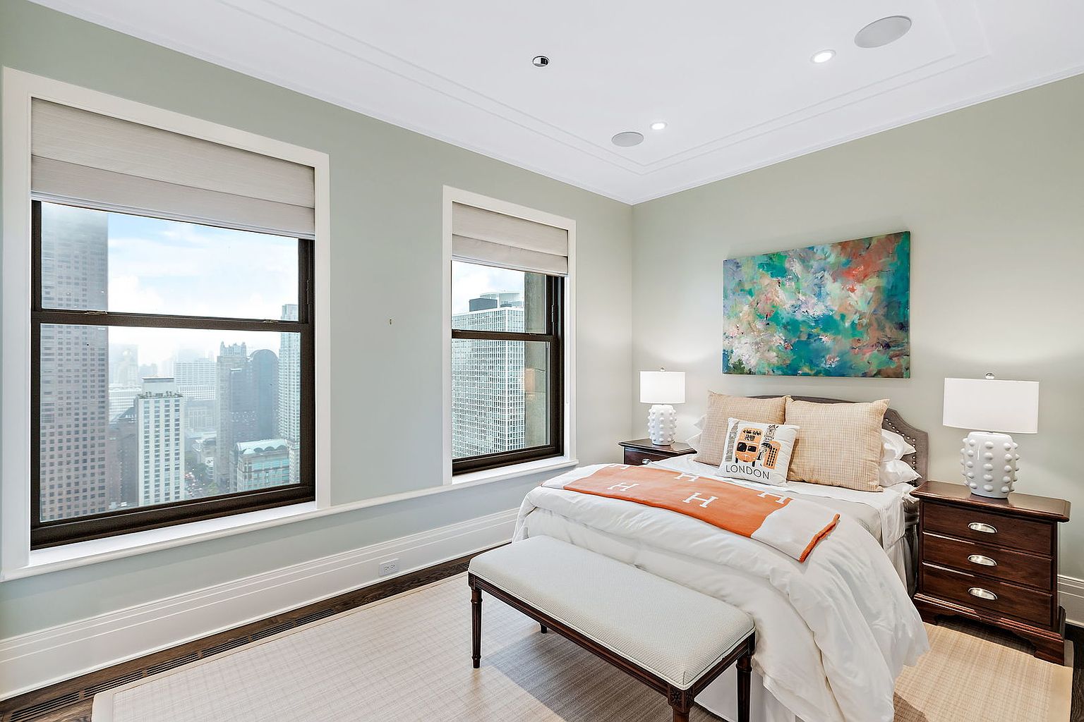 This is a bright and airy primary bedroom featuring two large windows with city views. The room is decorated in a soothing color palette with a large abstract painting above the bed. The bed is neatly made with an orange throw and decorative pillows, and there are matching nightstands on either side of the bed, each with a lamp.