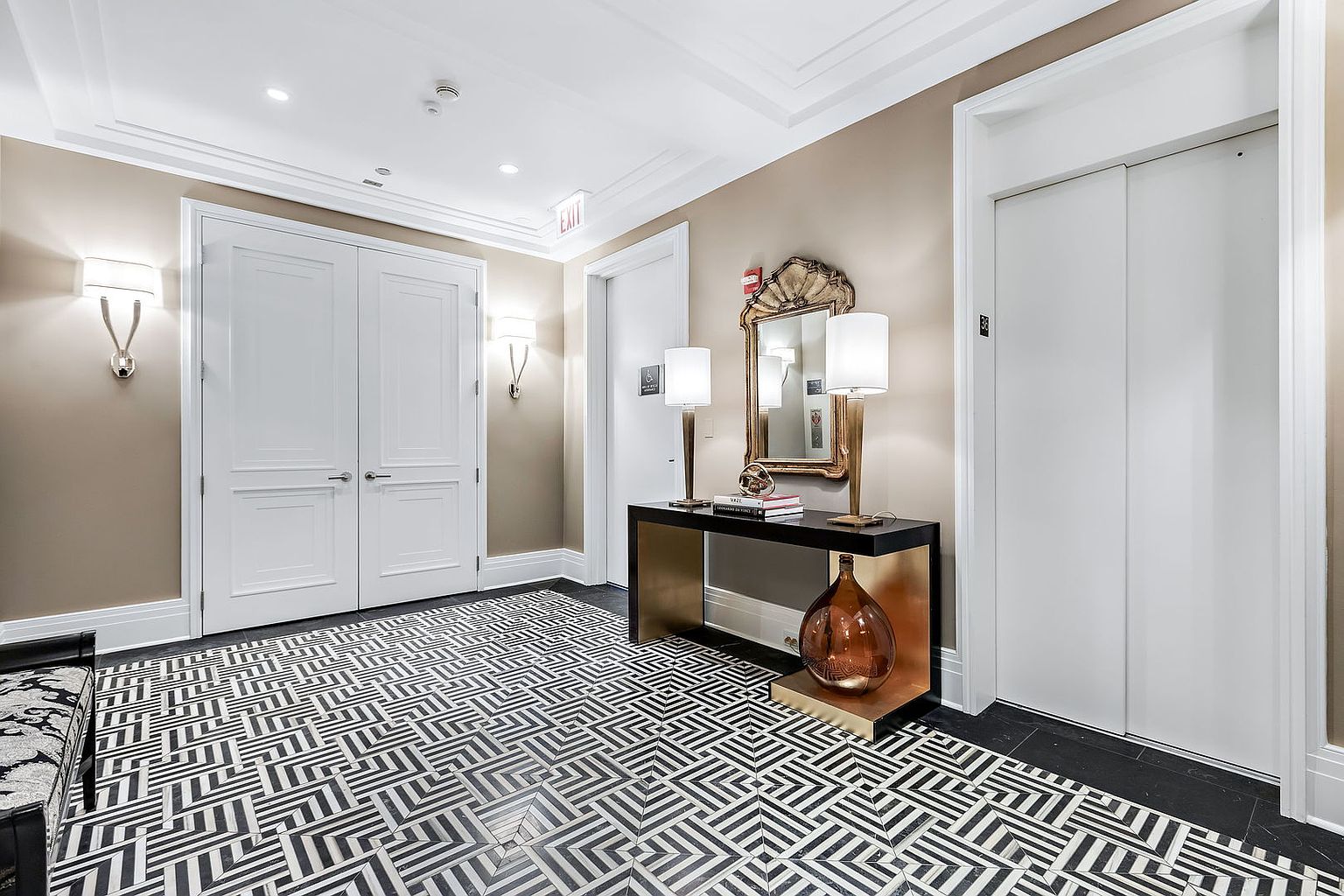 This interior shot showcases a stylish hallway with a striking black and white geometric tile floor. A console table with lamps and a decorative mirror adds elegance, while white doors and trim provide a clean contrast to the beige walls. The space is well-lit and exudes a sophisticated, modern aesthetic.