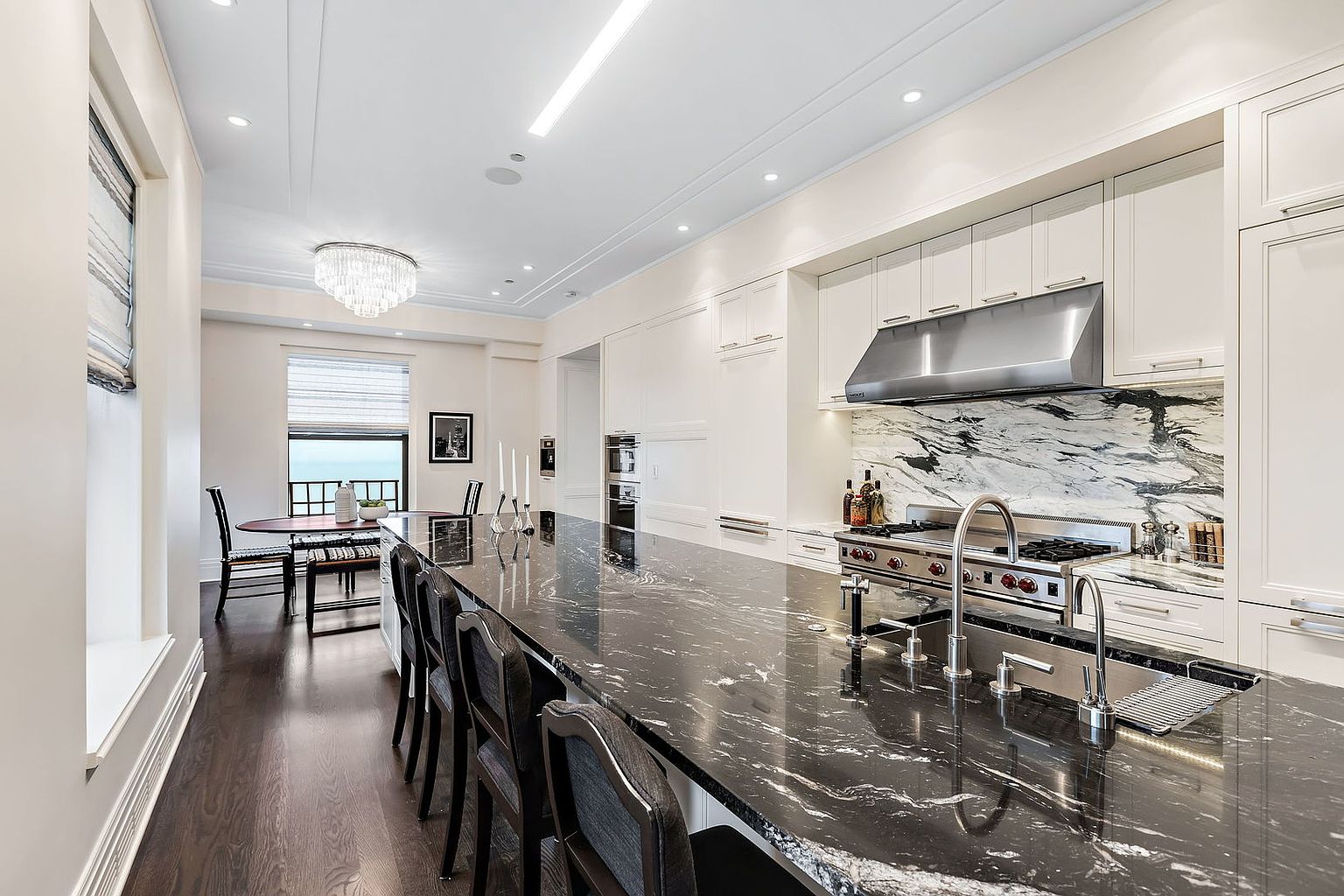 This is a bright, modern kitchen featuring white cabinetry, stainless steel appliances, and a large black marble island with seating. The kitchen also has a dining area with a chandelier and a view of the water. The perspective is from the side of the island, showcasing the length of the countertop and the appliances.