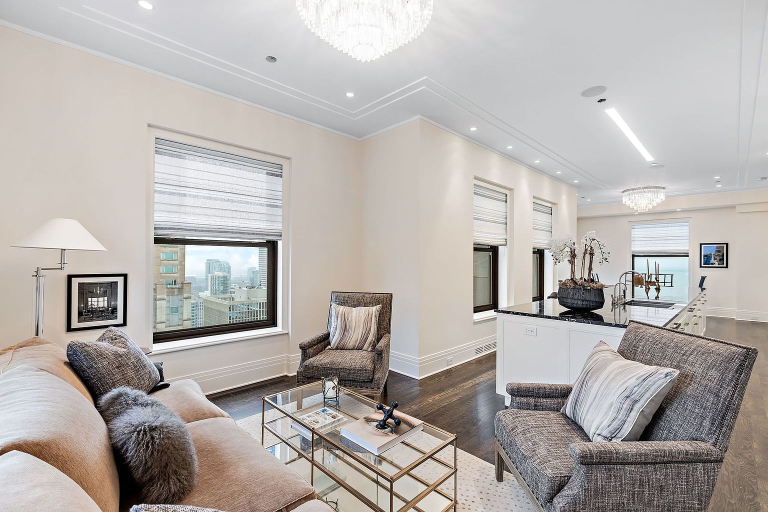 This is an interior shot of a luxurious living room and kitchen area. The living room features a comfortable sofa, stylish armchairs, and a glass coffee table, complemented by large windows offering city views. The kitchen area includes a sleek island with a dark countertop, creating a sophisticated and inviting space.