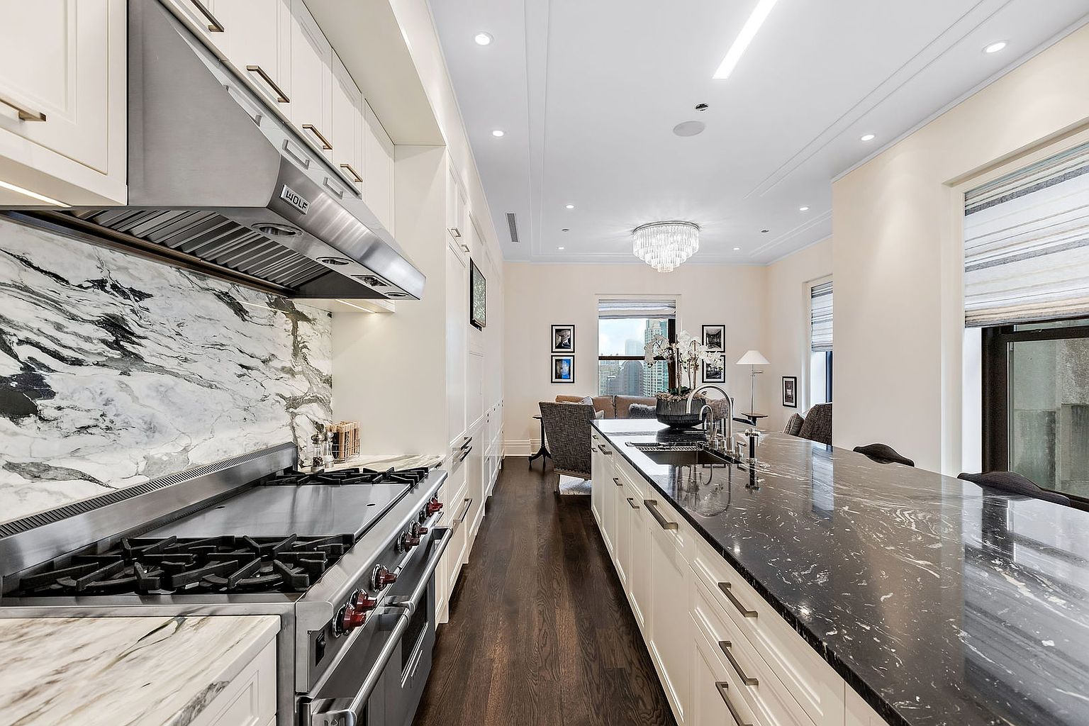 This is a modern kitchen with a long island featuring a dark countertop and white cabinetry. Stainless steel appliances, including a Wolf range and hood, are visible against a marble backsplash. The kitchen opens into a living area with a chandelier and city views, creating a seamless and luxurious living space.