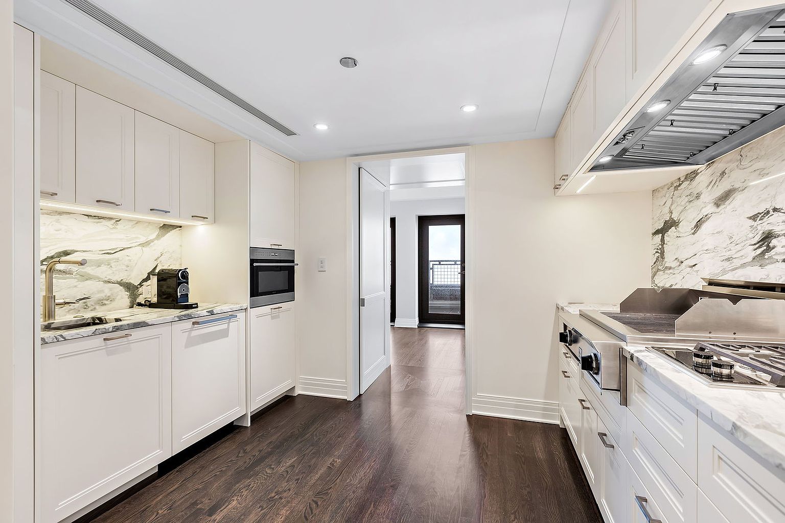 This is a well-lit kitchen featuring white cabinetry, marble countertops and backsplash, and stainless steel appliances. The dark hardwood flooring adds a touch of elegance, while the open doorway leads to a balcony, suggesting a seamless indoor-outdoor flow. The kitchen is clean, modern, and designed for both functionality and aesthetic appeal.