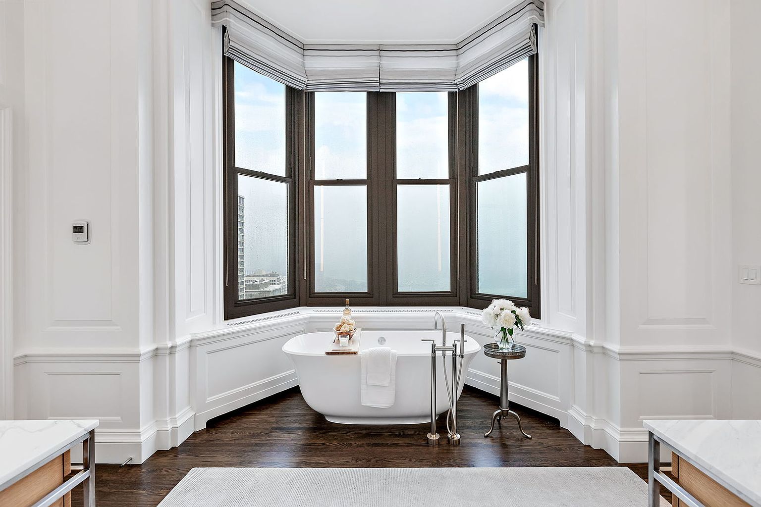 This is a luxurious primary bathroom featuring a freestanding bathtub positioned in front of a large bay window with striped blinds. The room is decorated in a classic style with white wainscoting, dark hardwood floors, and a light gray rug. A small side table with a vase of white flowers adds a touch of elegance.