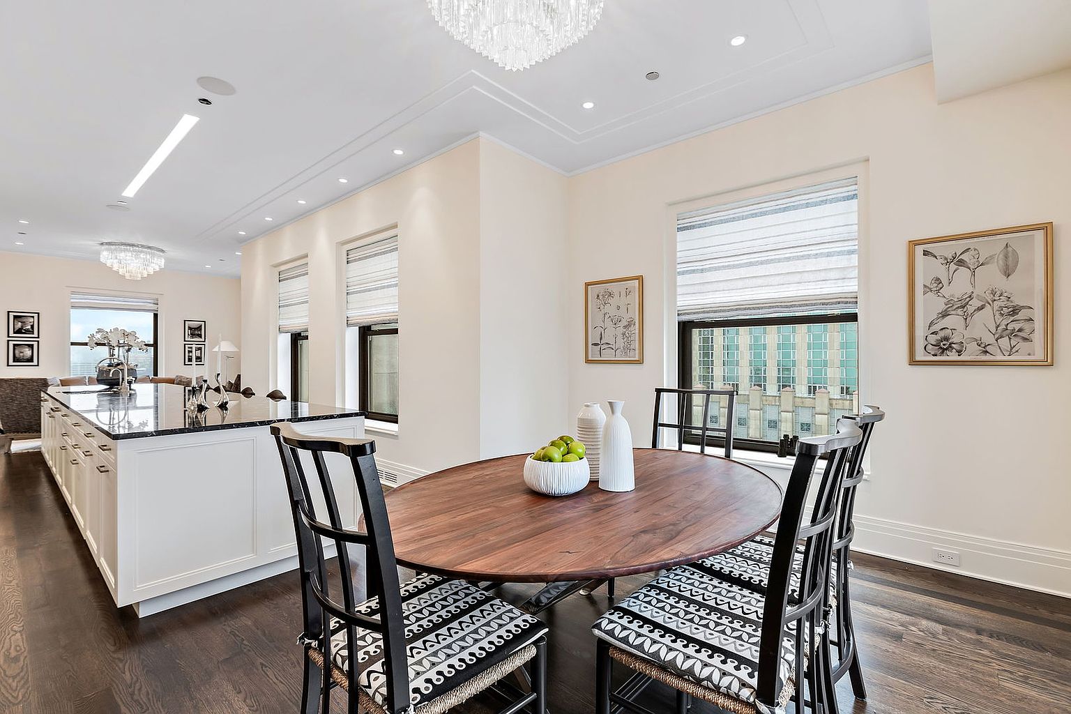 This interior shot showcases a dining area with a round wooden table surrounded by four black chairs with patterned cushions. The room features two windows with blinds, allowing natural light to filter in, and artwork adorns the walls. The dark wood flooring adds warmth and elegance to the space, creating an inviting atmosphere for dining.