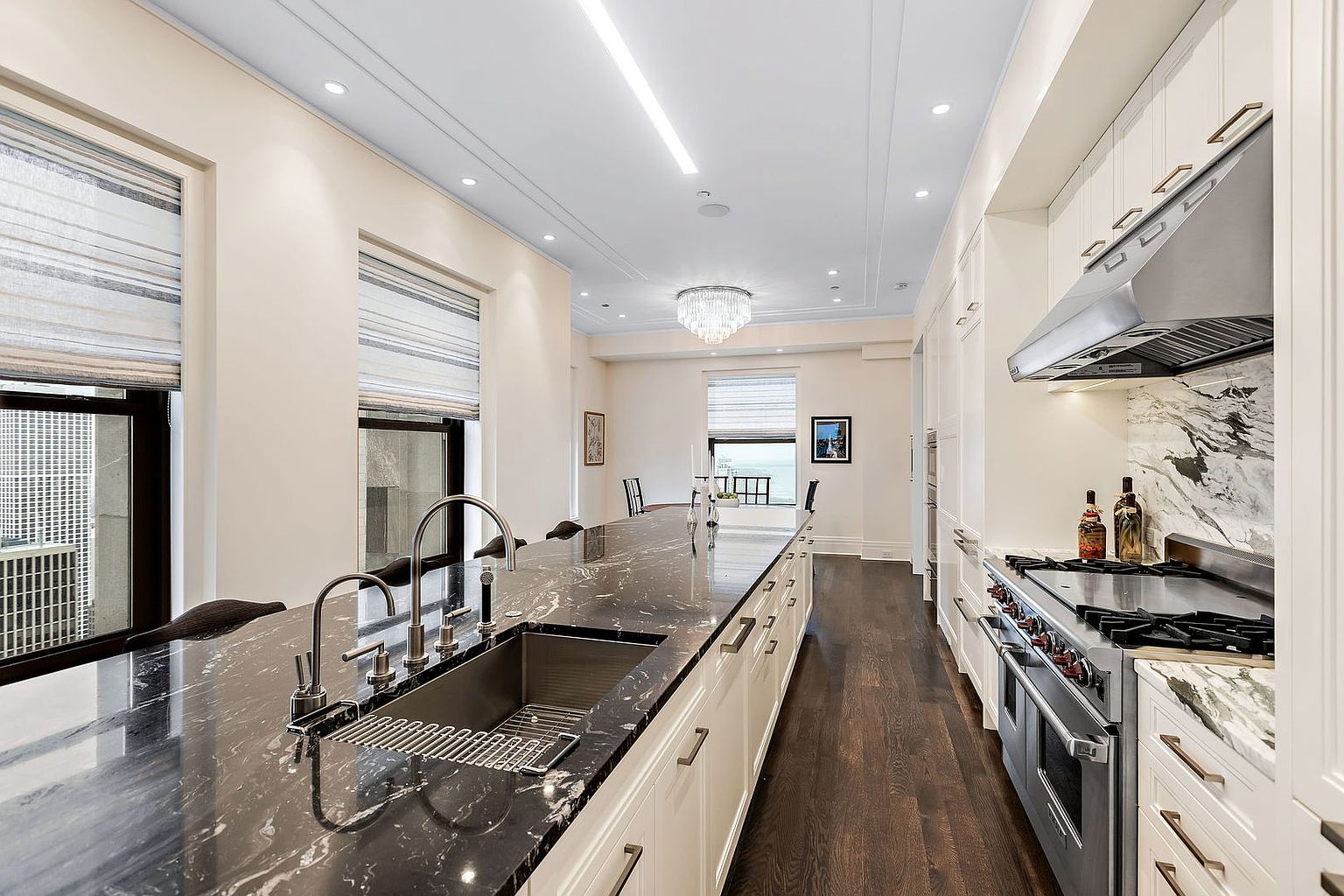 This is a bright and spacious kitchen featuring a long island with a dark marble countertop and stainless steel sink. The kitchen is equipped with high-end stainless steel appliances and white cabinetry, complemented by dark hardwood flooring. Large windows provide natural light, enhancing the elegant and modern design.