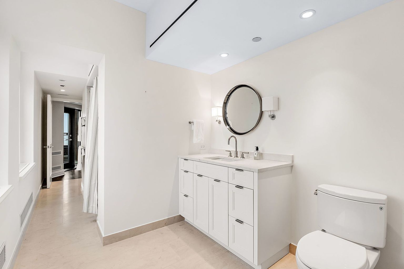 This is a well-lit bathroom featuring a white vanity with a marble countertop, a round mirror with a dark frame, and modern sconces. A toilet is visible to the right of the vanity. The bathroom has a clean and contemporary style with neutral-toned flooring, creating a bright and inviting space.