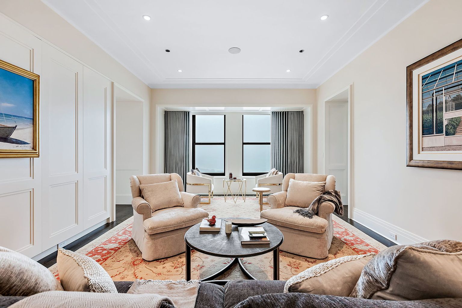 This is an interior shot of a living room featuring two plush armchairs facing each other, flanking a round coffee table. The room is decorated with neutral tones, complemented by an ornate rug and artwork on the walls. The space is well-lit with recessed lighting and natural light from the windows, creating a cozy and inviting atmosphere.