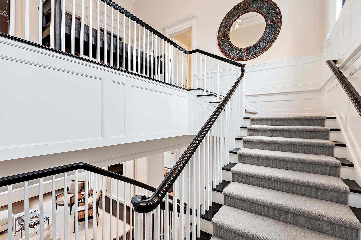 This image showcases an elegant interior staircase with a black handrail and white balusters. The stairs are carpeted in a neutral tone, and the walls feature white wainscoting. A decorative round mirror hangs on the wall, adding a touch of sophistication to the space.