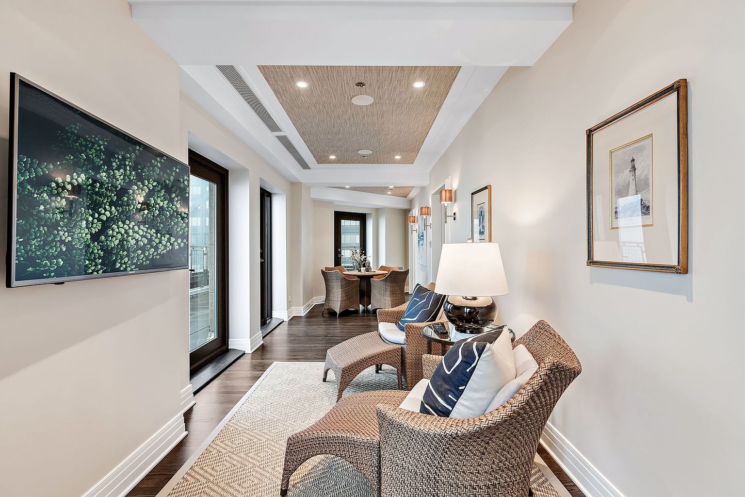 This interior shot showcases a well-lit hallway with elegant design elements. The hallway features a textured ceiling panel, recessed lighting, and artwork adorning the walls. Comfortable seating arrangements with wicker chairs and a patterned rug create an inviting and stylish transition space, while a large television adds a modern touch.
