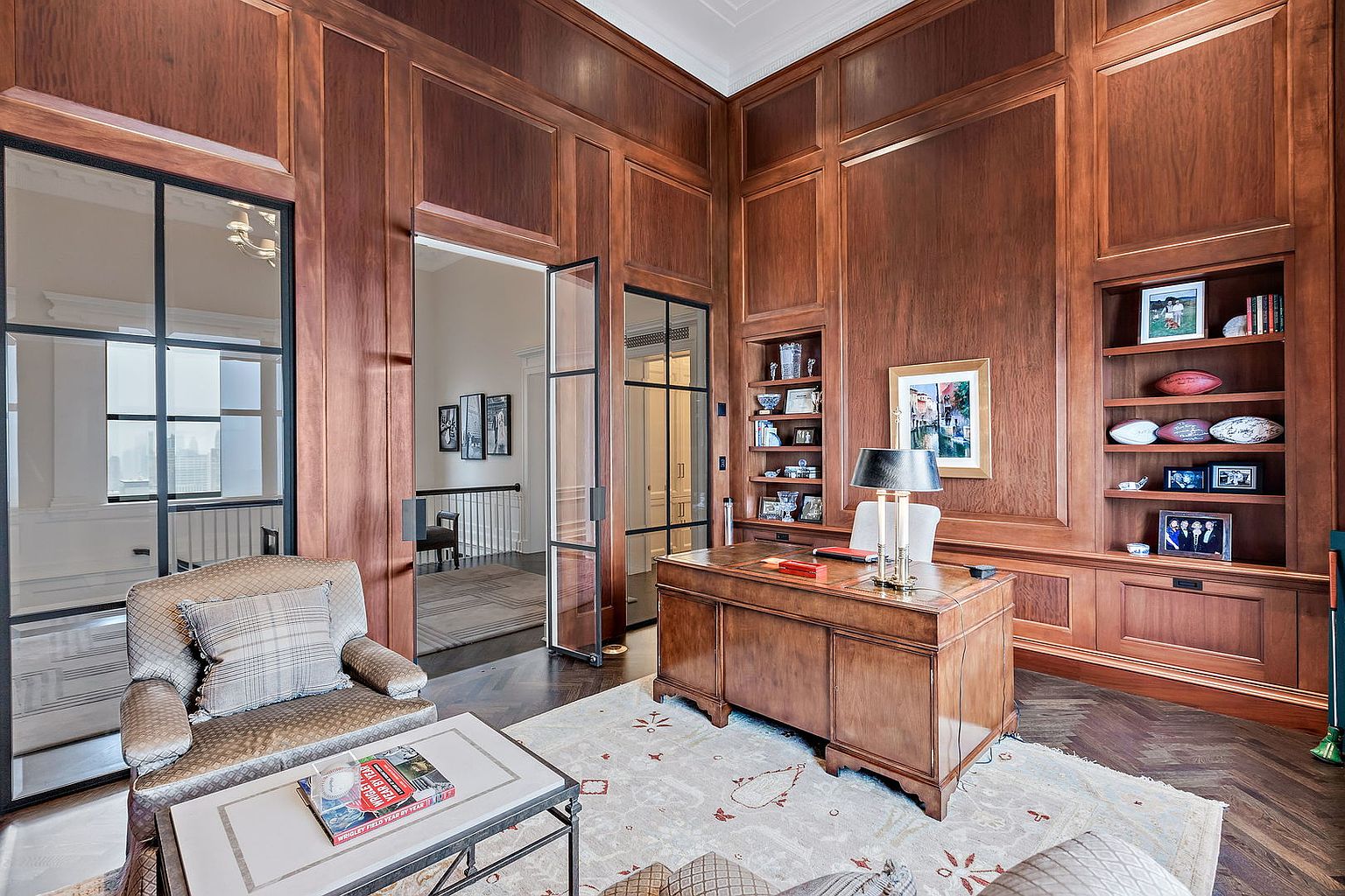 This is a luxurious home office featuring rich wood paneling, built-in bookshelves, and a stately wooden desk. The room is well-lit by a table lamp, and a comfortable armchair sits near a glass-paneled door, creating an inviting workspace. The overall impression is one of sophistication and classic elegance.