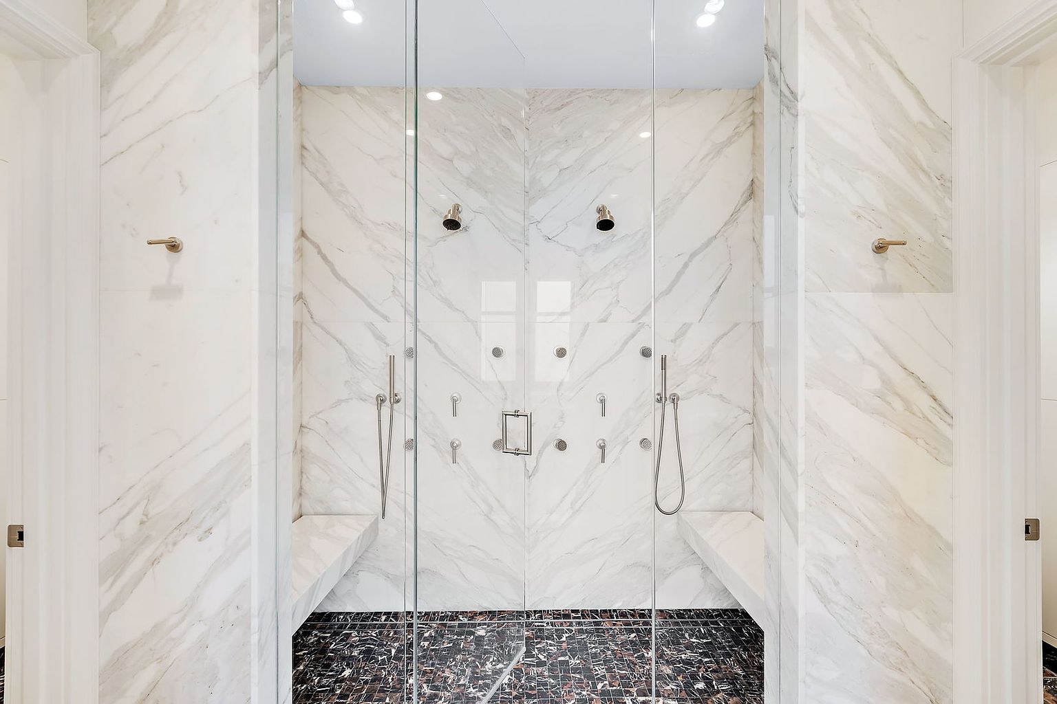 This is a luxurious primary bathroom featuring a large, glass-enclosed shower with dual shower heads and body jets. The walls are clad in elegant marble, complemented by a dark, patterned tile floor. Built-in marble benches offer seating within the shower, enhancing the spa-like atmosphere.