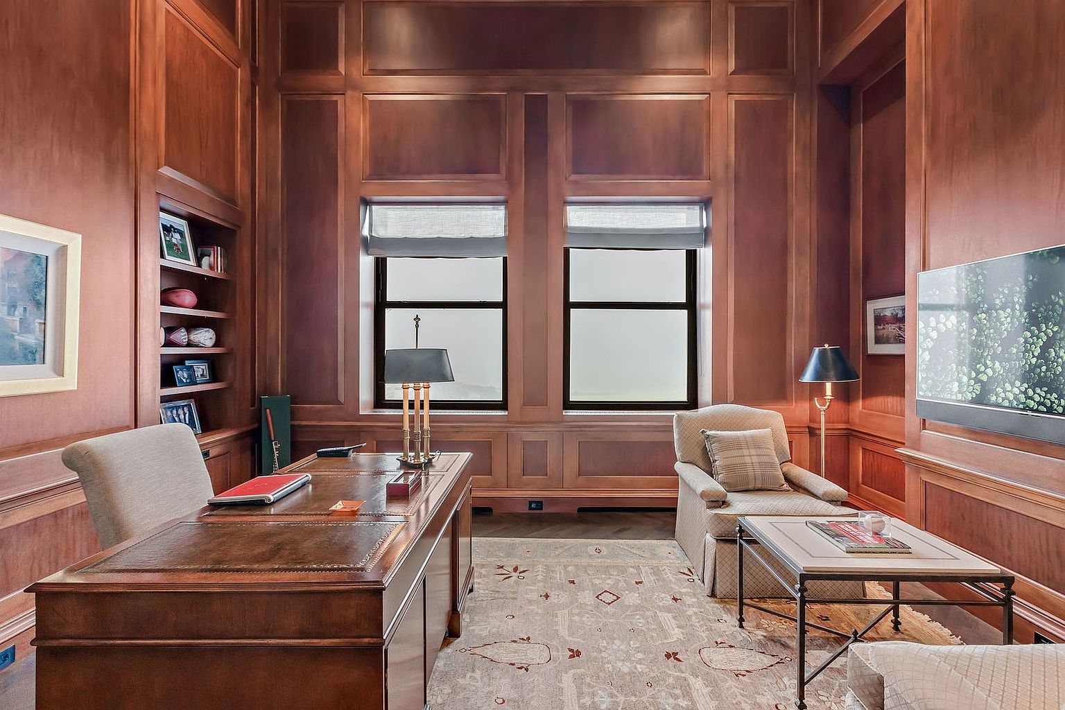 This is a classic and sophisticated home office featuring rich wood paneling and built-in bookshelves. A large, leather-topped desk dominates the space, complemented by a comfortable armchair and a patterned area rug. Natural light filters through the windows, creating a warm and inviting atmosphere, while a television is mounted on the wall.
