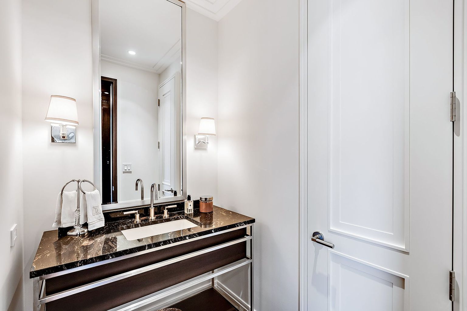 This is a well-lit guest bathroom featuring a dark marble countertop vanity with a chrome frame and a rectangular sink. A large mirror hangs above the vanity, flanked by sconce lighting. A white paneled door is visible to the right, contributing to the clean and modern aesthetic of the space.