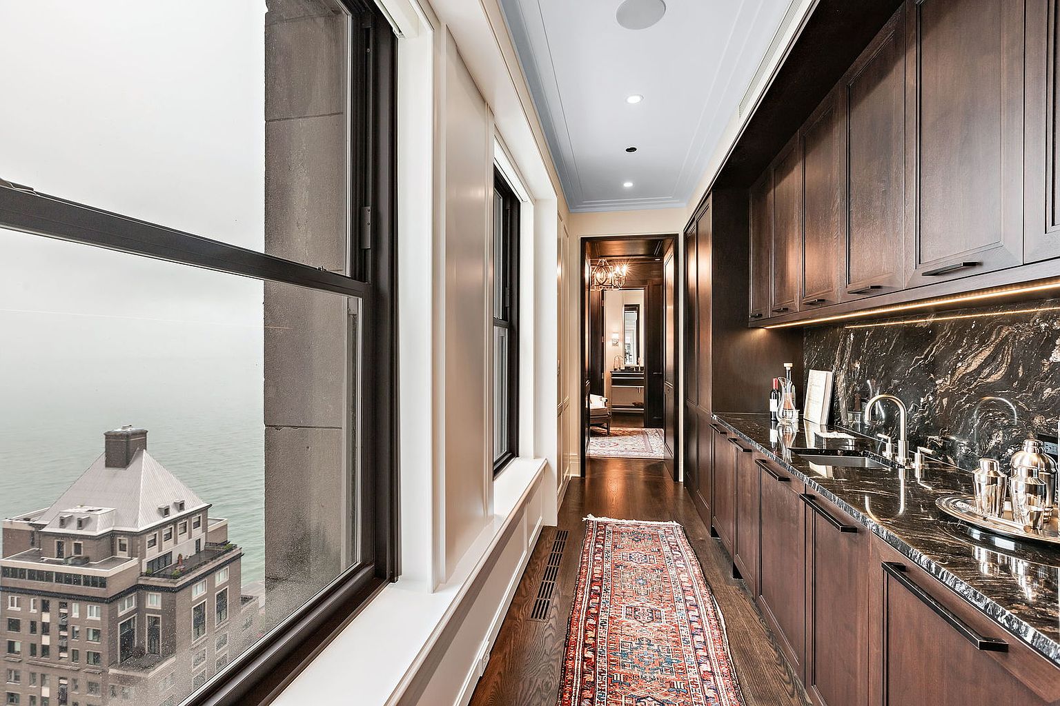 This interior shot showcases a hallway featuring dark wood cabinetry and countertops with a marble backsplash. A long, ornate rug runs the length of the hallway, leading towards a doorway with a glimpse into another room. Large windows offer a view of a nearby building and the water, adding natural light to the space.