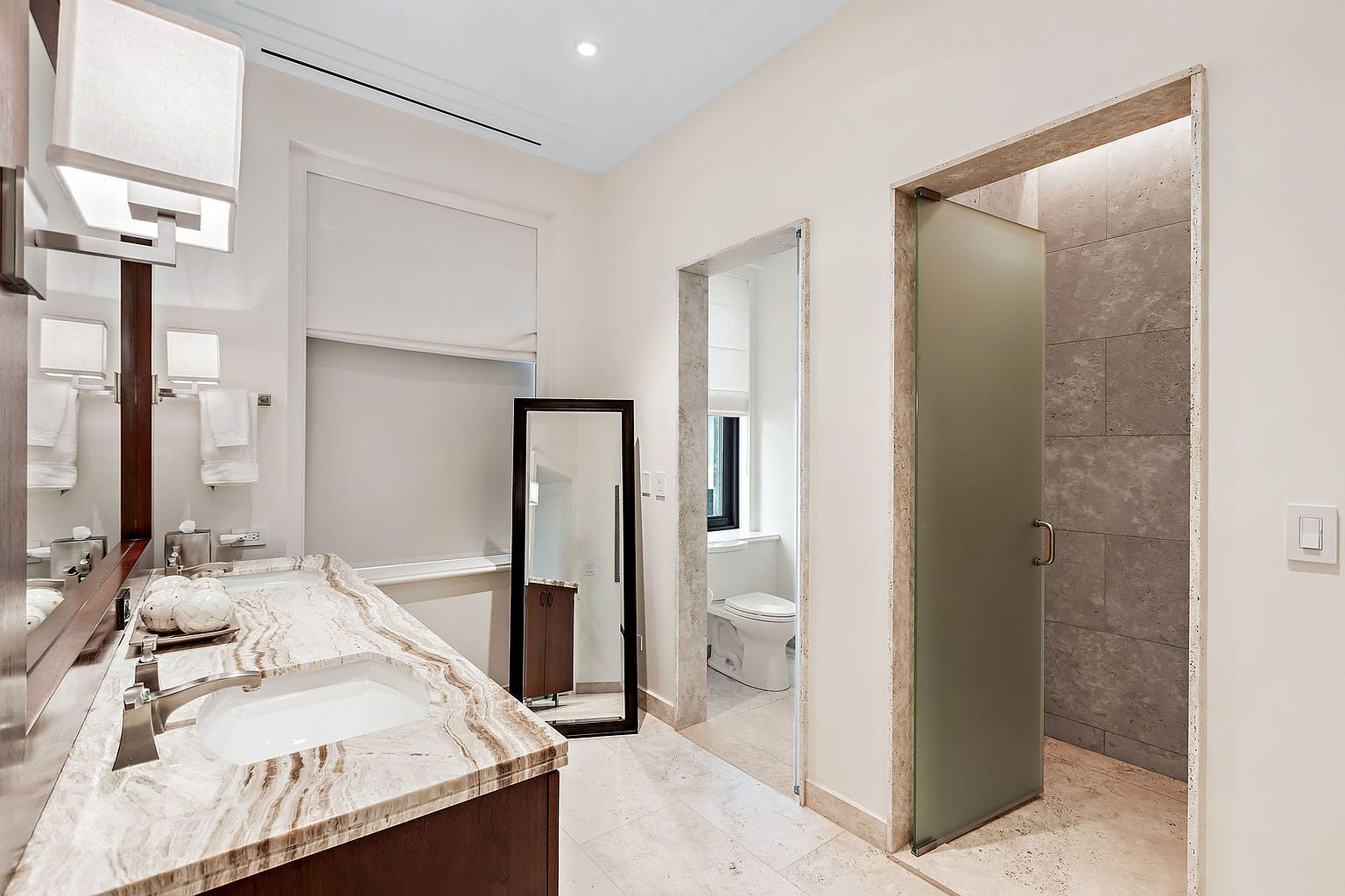 This is a primary bathroom featuring a double sink vanity with a marble countertop and dark wood cabinetry. A large mirror and modern lighting fixtures are above the vanity. The bathroom also includes a separate toilet room and a shower room with a frosted glass door, both accessible through doorways. The overall style is modern and luxurious.