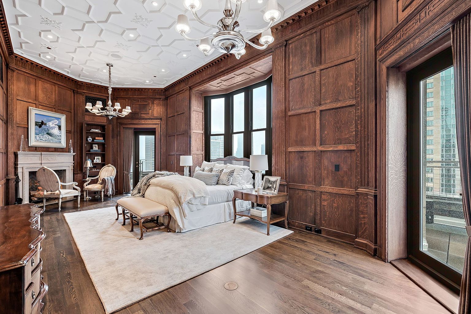 This is a luxurious primary bedroom featuring rich wood paneling, a coffered ceiling with intricate detailing, and a large bay window offering city views. The room is furnished with a plush bed, elegant seating, and a fireplace, creating a warm and inviting atmosphere. The perspective is a wide shot, capturing the grandeur and spaciousness of the room.