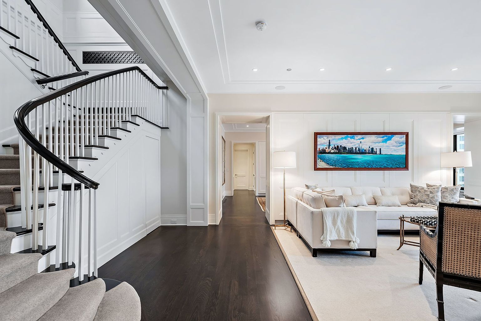 This interior shot showcases a grand hallway with a staircase and a glimpse into a living area. The staircase features a dark wood handrail contrasting with white spindles, leading to the upper level. The hallway transitions into a living space with a comfortable sofa and artwork, creating an inviting and luxurious atmosphere.