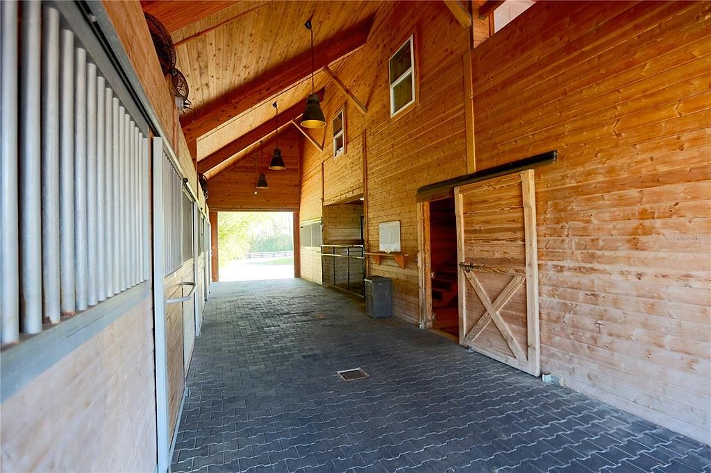 This image showcases the interior of a well-maintained barn, featuring wooden walls and a dark brick floor. Stalls line one side, while a partially open wooden door reveals a glimpse of the interior space. The barn is well-lit with hanging lights, creating a warm and inviting atmosphere.