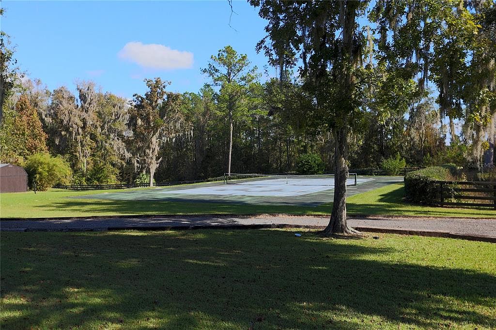 This image showcases a well-maintained yard with a tennis court as a prominent feature. The landscape includes mature trees, some draped with Spanish moss, adding a touch of Southern charm. The scene is bathed in natural light, creating a serene and inviting atmosphere, perfect for outdoor activities and relaxation.
