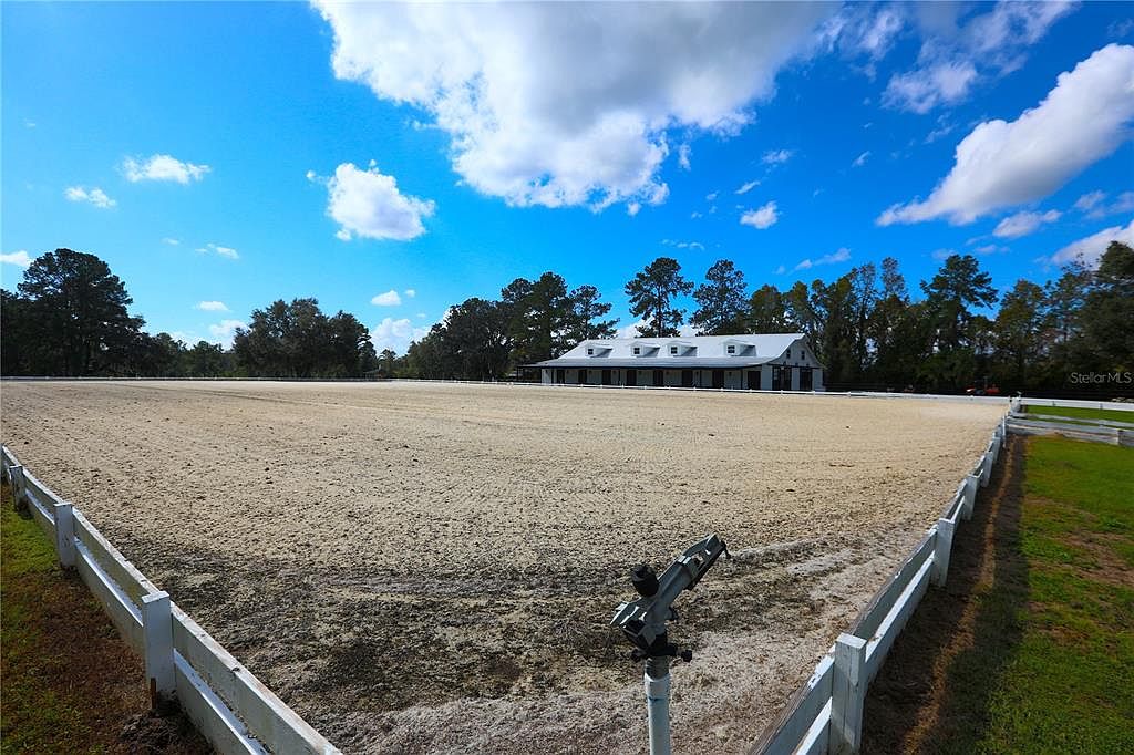 The image showcases a large, well-maintained equestrian arena with a white fence surrounding the perimeter. In the background, a stable or barn with a white roof is visible, set against a backdrop of trees and a bright blue sky with scattered clouds. The arena's sandy surface appears smooth and ready for use, creating an inviting space for equestrian activities.