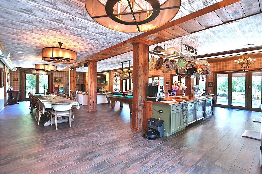 This is a spacious, open-concept kitchen and dining area featuring a rustic design with wooden beams and flooring. The kitchen island is equipped with modern appliances and a unique pot rack, while the dining area includes a large table set for a meal. The room is well-lit with a combination of natural light and decorative chandeliers, creating a warm and inviting atmosphere.