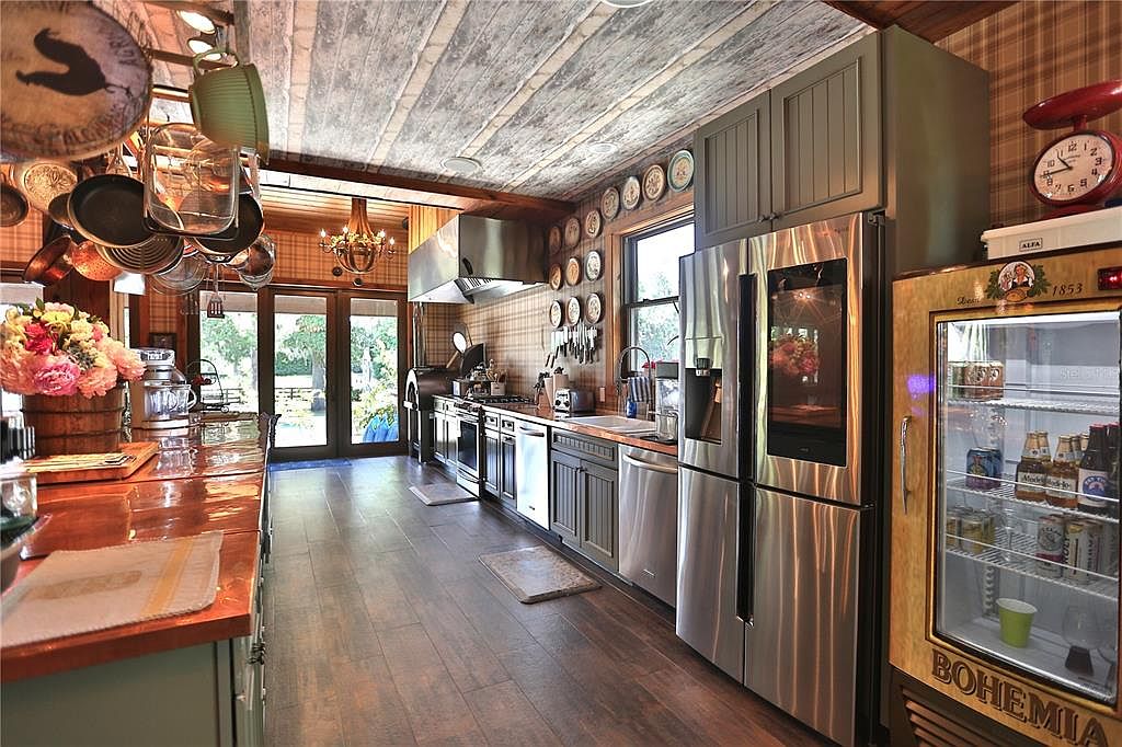 This is a warm and inviting kitchen featuring a unique blend of rustic and modern elements. The kitchen boasts stainless steel appliances, custom cabinetry, and a striking wood-planked ceiling. Decorative plates and hanging cookware add to the charm, while the open layout and natural light create a welcoming atmosphere.