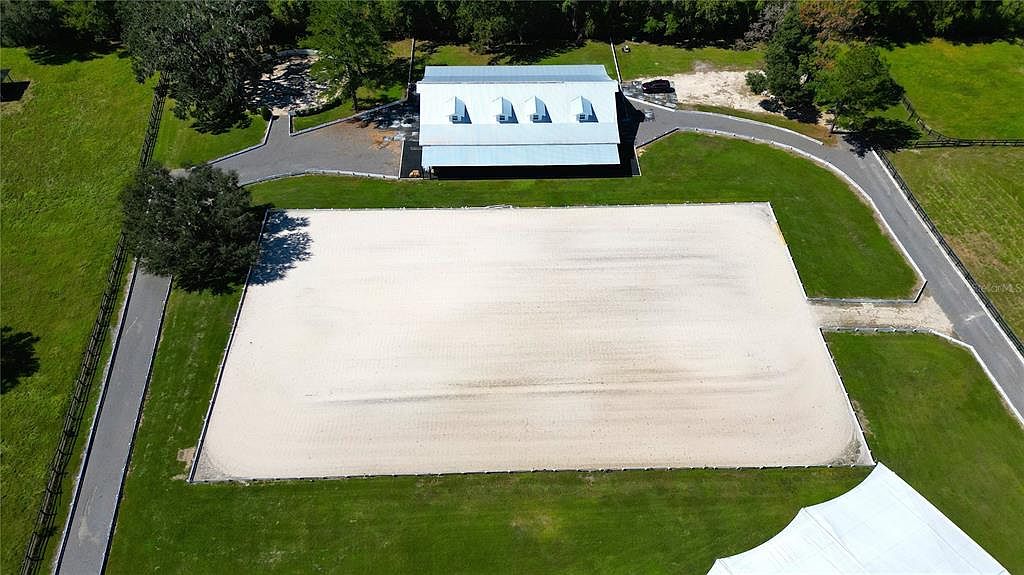 This aerial shot showcases a large equestrian arena with a pristine sand surface, bordered by lush green lawns and mature trees. A stable building with a metal roof is visible in the background, along with paved driveways leading to the property. The overall impression is one of a well-maintained and spacious equestrian estate.
