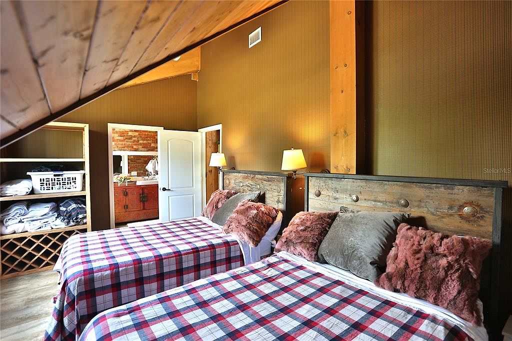 This is an interior shot of a guest bedroom featuring two twin beds with plaid bedding, rustic wooden headboards, and decorative pillows. The room has a warm, inviting ambiance with wooden beams and a view into an adjacent bathroom with brick accents. A wooden shelving unit provides storage, enhancing the room's functionality.