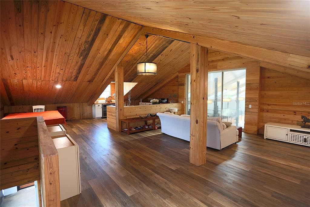 This is an interior shot of a living room with a rustic, cabin-like feel. The room features wood paneling on the walls and ceiling, hardwood floors, and a light-colored sofa. A partial kitchen area is visible in the background, and natural light streams in through a large window, creating a warm and inviting atmosphere.