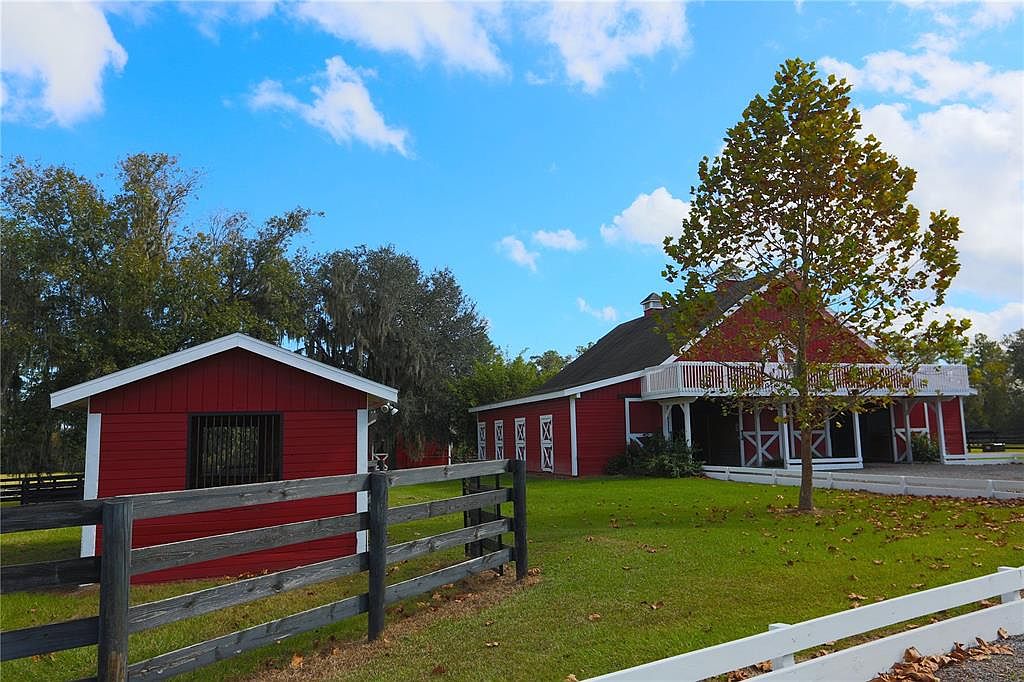 This image showcases a well-maintained yard with two red barns featuring white trim. A wooden fence runs along the front of the property, adding to the rustic charm. The sky is blue with scattered clouds, creating a picturesque and inviting scene.