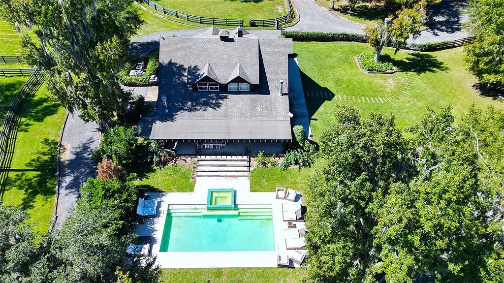 This aerial shot showcases a beautiful property featuring a large house with a gray roof, a swimming pool with a jacuzzi, and a well-maintained yard. The house has a covered patio area with outdoor seating, and the surrounding landscape includes mature trees and fencing, suggesting a private and spacious estate. The perspective is high-angle, providing a comprehensive view of the property's layout and features.