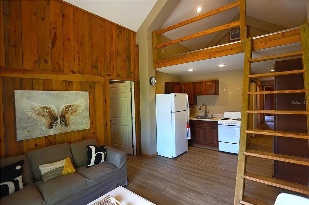 This interior shot showcases a cozy living space with wood-paneled walls and a comfortable gray sofa adorned with decorative pillows. The room features a unique loft area accessible by a wooden ladder, adding architectural interest. A compact kitchen area is visible in the background, creating an open-concept living space.