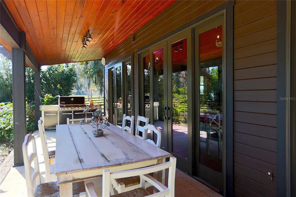 This image showcases a covered patio area, perfect for outdoor dining and entertaining. A long wooden table with white chairs sits prominently, with a stainless steel grill visible in the background. The patio features a wooden ceiling and sliding glass doors, creating a seamless transition between indoor and outdoor living spaces.
