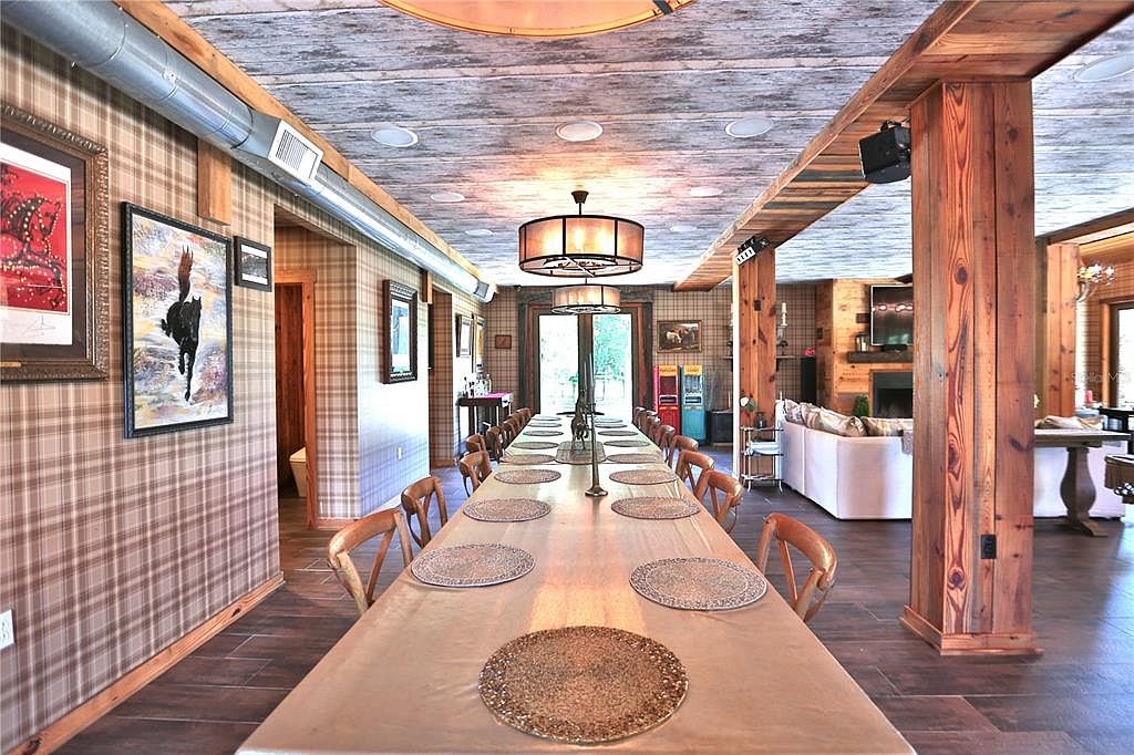 This is an interior shot of a dining room featuring a long table set for many guests. The room has a rustic yet elegant style, with plaid wallpaper, wooden beams, and modern light fixtures. The perspective is from the end of the table, looking towards a doorway and a glimpse of a living area, creating a sense of depth and spaciousness.