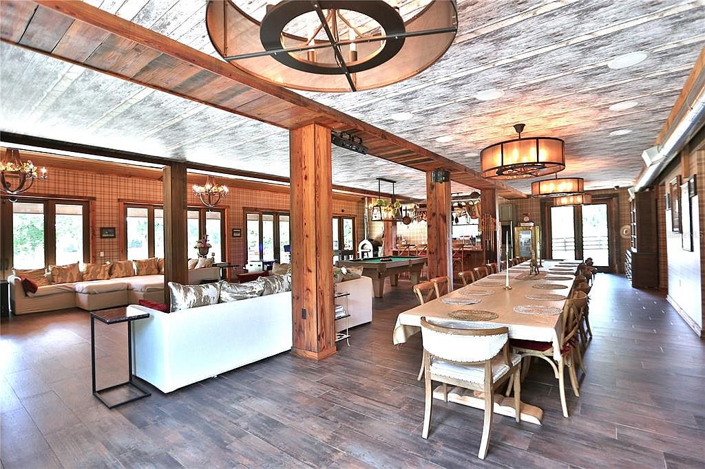 This is a spacious living room featuring a large dining table with multiple chairs, a pool table, and a comfortable seating area with sofas. The room is well-lit with a combination of natural light from the windows and artificial lighting from chandeliers and ceiling fixtures. The flooring is wood-like tile, and the walls are covered in a plaid pattern, creating a warm and inviting atmosphere.