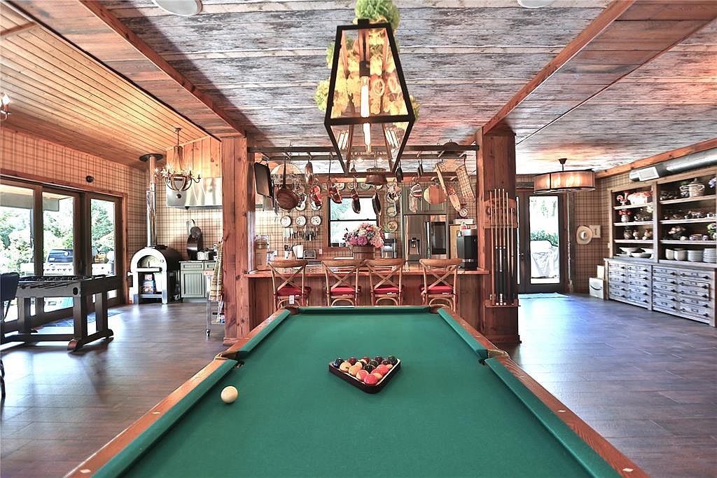 This interior shot showcases a spacious living room with a rustic yet elegant design. A pool table takes center stage, complemented by a bar area with seating and a unique overhead lighting fixture. The room features wood paneling, a decorative ceiling, and various decorative elements, creating a warm and inviting atmosphere.