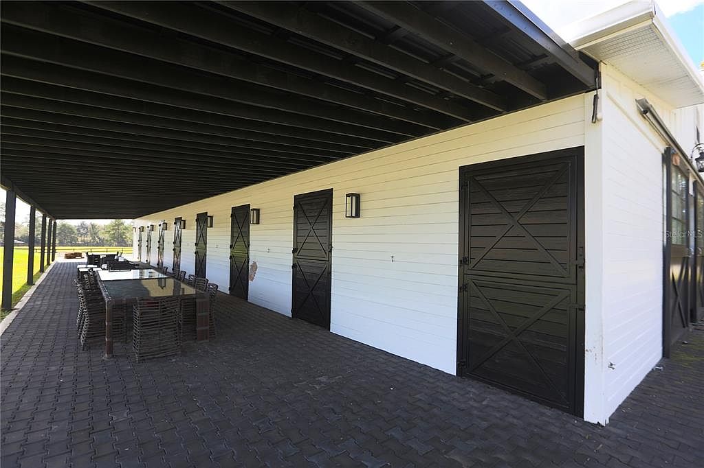 This image showcases a covered patio area with a long dining table and chairs, adjacent to a building with white siding and black doors. The patio is paved with dark bricks, and the black ceiling provides shade. The scene suggests an outdoor dining or entertainment space, perfect for gatherings.