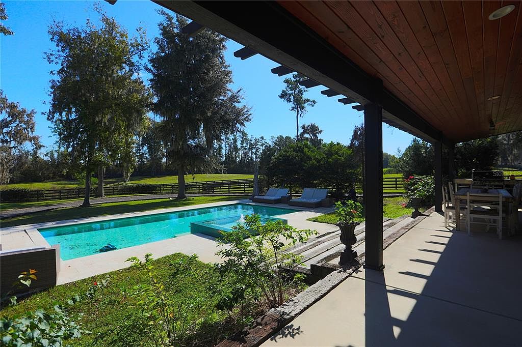 This image showcases a luxurious outdoor space featuring a rectangular swimming pool with a built-in spa. The pool area is surrounded by a well-manicured lawn, mature trees, and a wooden fence, creating a serene and private setting. A covered patio with outdoor seating extends from the house, offering a perfect spot for relaxation and entertainment.