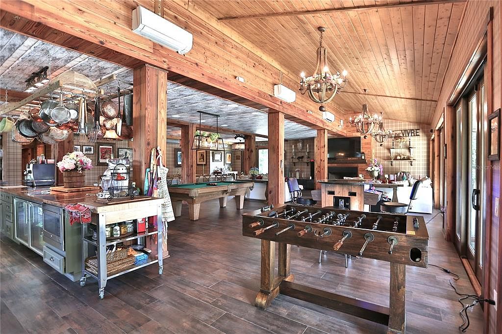 This expansive living room features a rustic design with wood-paneled walls and ceilings, complemented by dark wood flooring. The space includes a kitchen area with hanging pots and pans, a pool table, and a foosball table, creating a versatile entertainment zone. Chandeliers provide warm lighting, enhancing the room's inviting atmosphere.
