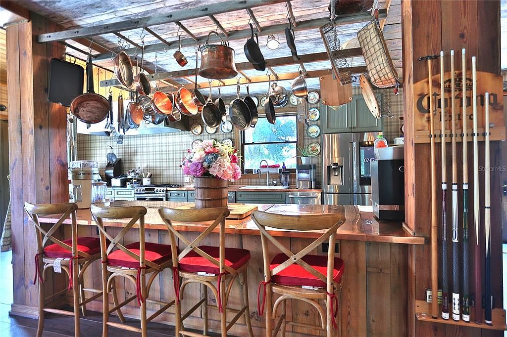 This is a warm and inviting kitchen featuring rustic wood paneling and a unique pot rack suspended from the ceiling. The kitchen island is lined with wooden bar stools with red cushions, and a Corona-themed pool cue rack adds a touch of character. The overall impression is cozy and charming, perfect for a country-style home.