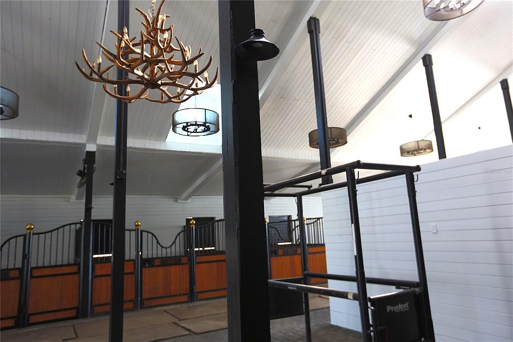 The image showcases the interior of a horse stable, featuring wooden stalls with black metal bars and gold accents. A unique antler chandelier hangs from the white, vaulted ceiling, complemented by modern light fixtures. The space is well-lit and appears clean, suggesting a high-end equestrian facility.