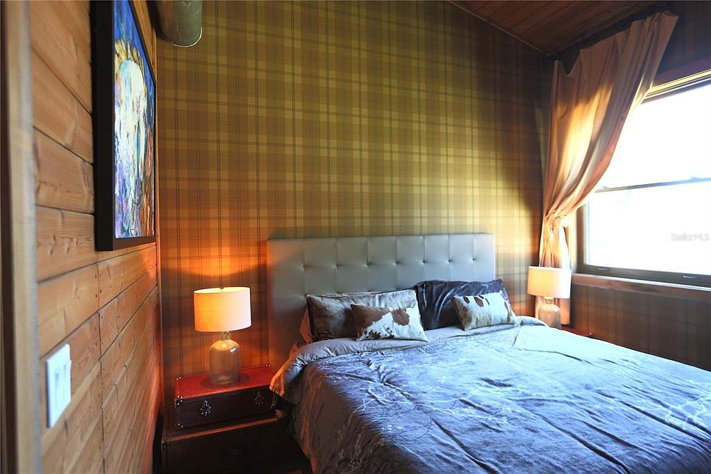 This is an interior shot of a bedroom featuring a bed with a gray headboard and blue bedding. The walls are covered in a plaid wallpaper, and there is a window with curtains on the right side of the image. A painting hangs on the wood-paneled wall to the left, and two lamps sit on either side of the bed on small tables.