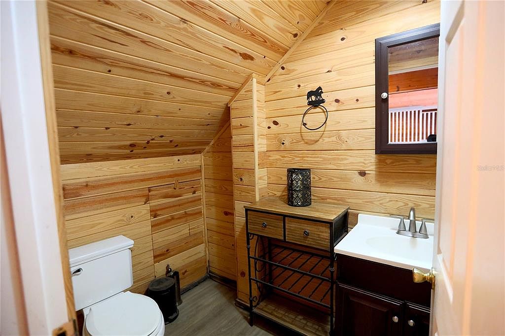 This is a cozy bathroom featuring wood-paneled walls and ceiling, giving it a rustic cabin feel. The bathroom includes a toilet, a vanity with a white sink and dark wood cabinet, and a decorative mirror. A small cabinet with drawers and shelves provides additional storage, and a whimsical horse-shaped towel ring adds a touch of charm.