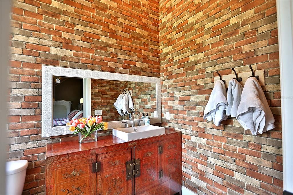 This is a bathroom featuring brick walls, a wooden vanity with a white sink, and a large mirror. Three towels hang on hooks on the wall. The style is rustic and unique, creating a warm and inviting atmosphere.