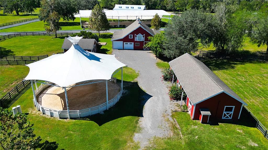 This aerial shot showcases a well-maintained equestrian estate. The property features a large covered riding arena, a red barn with white trim, and a long stable building, all connected by a gravel driveway. Lush green lawns and mature trees surround the buildings, creating a picturesque and serene setting.