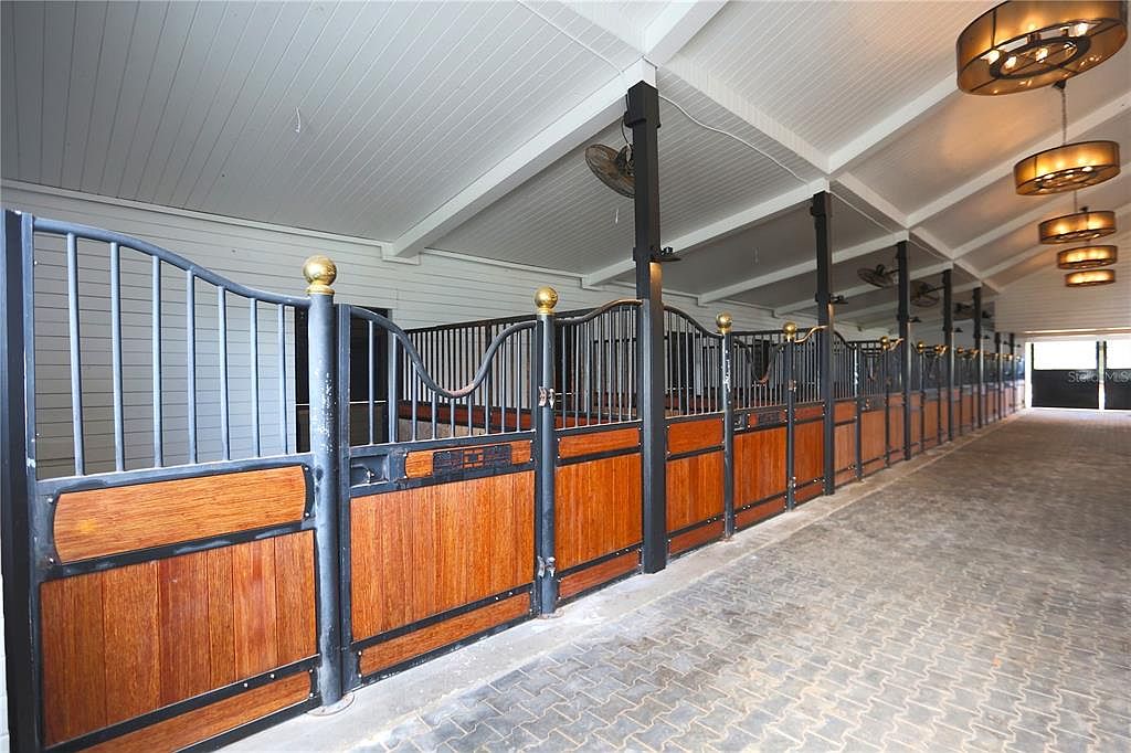 The image showcases an interior stable area with multiple horse stalls. The stalls feature wooden lower sections and metal barred upper sections, topped with decorative gold spheres. Overhead, there are modern light fixtures and ceiling fans, creating a well-lit and ventilated space. The floor is paved with interlocking bricks.