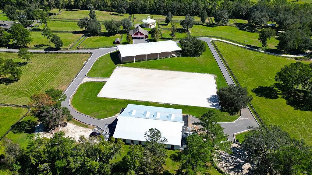 This aerial shot showcases a sprawling estate with lush green lawns, mature trees, and well-maintained landscaping. The property features a large riding arena, a covered training area, and several outbuildings, suggesting equestrian facilities. A paved driveway winds through the grounds, connecting the various structures and highlighting the property's spacious layout.
